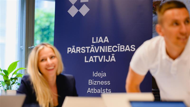 Foto no LIAA pasākuma, kurā redzams organizācijas nosaukums un otrajā plānā divas personas
