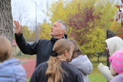 Bērni (ar skatu no mugurpuses) laukā pie koka stumbra klausās pasniedzēja stāstījumu