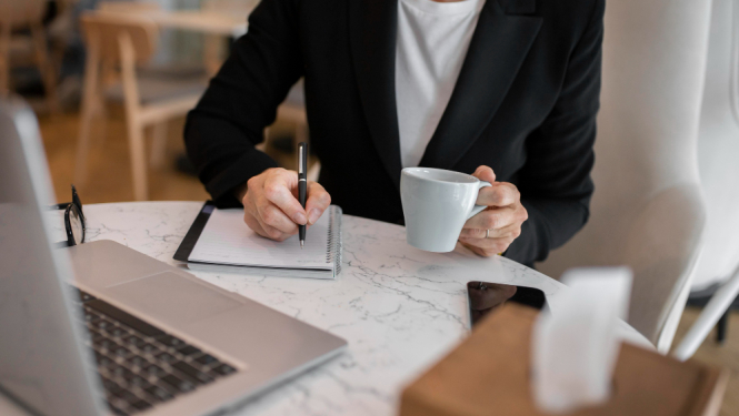 Sieviete ar kafijas krūzi raksta plānotājā, priekšplānā portatīvais dators