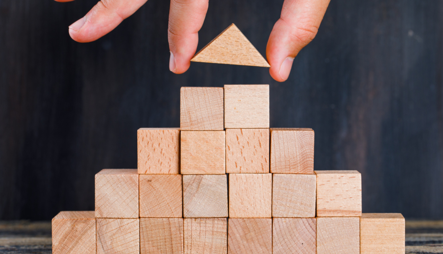 Koka klucīšu piramīda uz tumša fona. Roka uzliek pēdējo klucīti - slīpu jumtu.